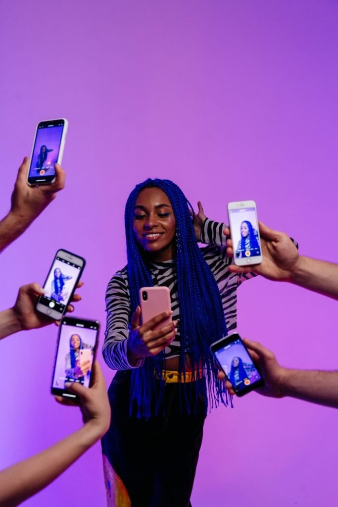 Portrait of a stylish woman with blue hair taking selfies surrounded by smartphones in a studio setting.